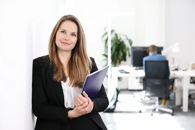 Une jeune agente immobilière se tient au milieu d'un bureau lumineux, regardant directement l'appareil photo avec confiance. À sa posture, on devine un grand professionnalisme et une attention toute particulière.